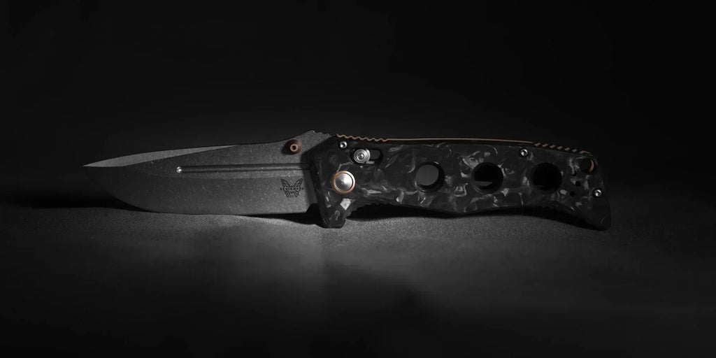 Benchmade Adamas in marbled carbon fiber with bronze hardware, open and photographed low-angle against a dark backdrop, emphasizing the blade edge and handle contours.