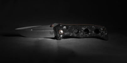 Benchmade Adamas in marbled carbon fiber with bronze hardware, open and photographed low-angle against a dark backdrop, emphasizing the blade edge and handle contours.