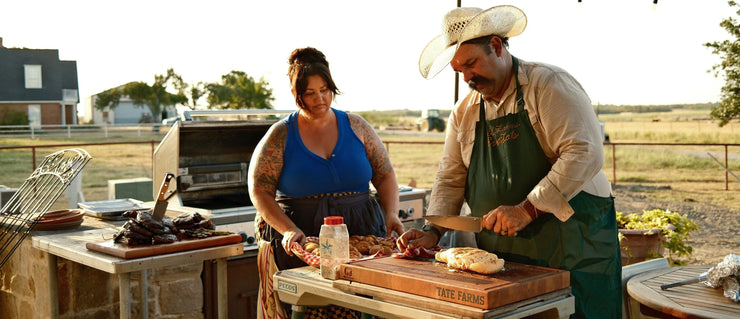 Melissa and Evan Tate preparing steak at an outdoor kitchen on Tate Farms, with Evan slicing meat on a Tate Farms cutting board