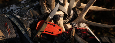 Sharp folding hunting knife with orange sheath rests on deer antlers and camo gear, ready for the field.