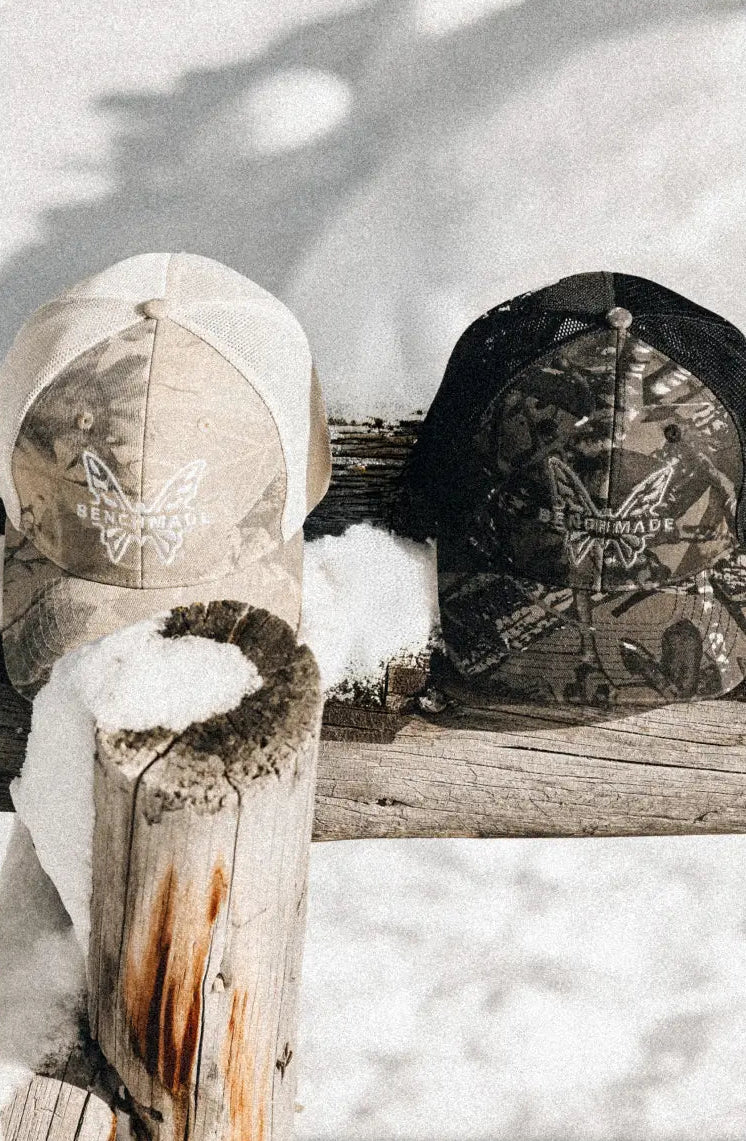 Two camouflage trucker-style baseball caps with a butterfly logo and 'BENCHMADE' text, displayed outdoors on a snowy wooden bench with a rustic log in the foreground.